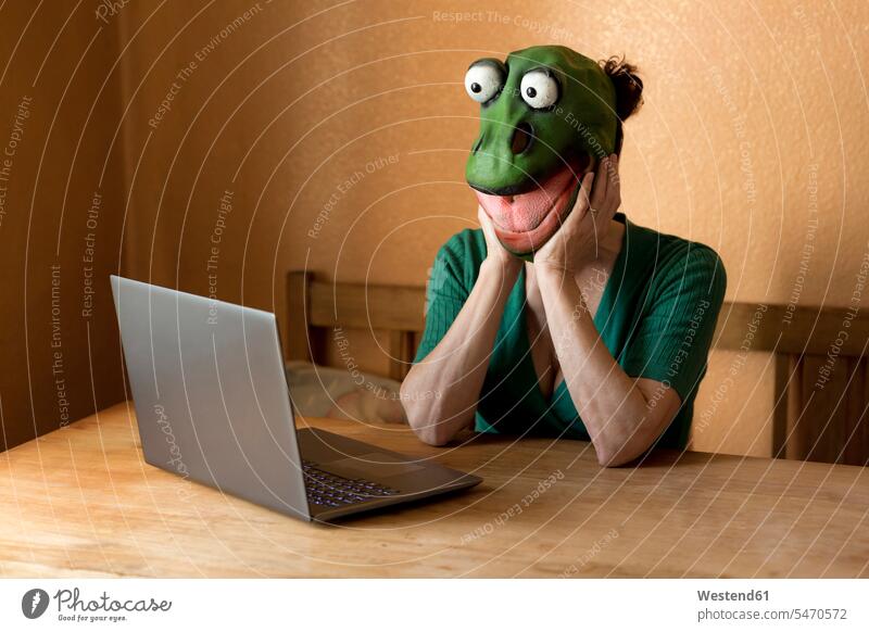 Playful mature woman wearing frog mask with laptop on table sitting at home during curfew color image colour image Germany indoors indoor shot indoor shots