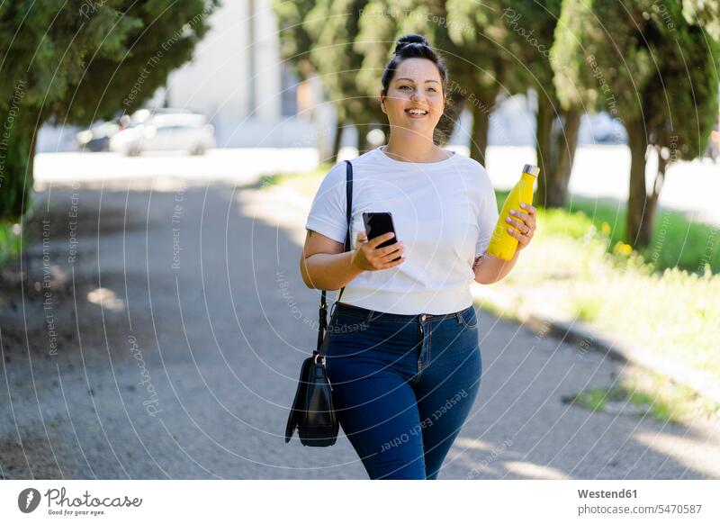 Smiling curvy young woman with mobile phone and bottle in a public park human human being human beings humans person persons caucasian appearance