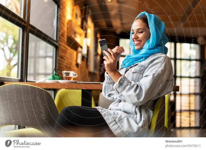 Young woman wearing turquoise hijab and using smartphone in a cafe windows head cloth head cloths head scarf head scarves headscarves telecommunication phones