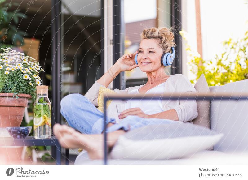 Portrait of smiling mature woman sitting on terrace listening music with headphones Bottles Glass Bottles Crockery Tableware Drinking Glass Drinking Glasses
