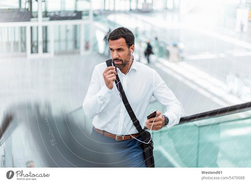 Businessman using smartphone, earphones, on escalator portrait portraits men males Business man Businessmen Business men Business Trip executive travel