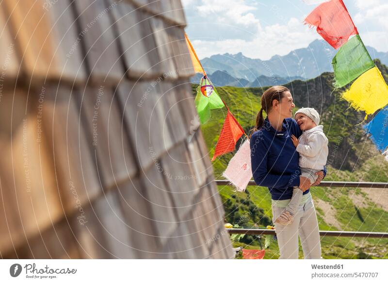 Germany, Bavaria, Oberstdorf, happy mother carrying little daughter on a mountain hut surrounded by pennants daughters streamer streamers mountain shelter