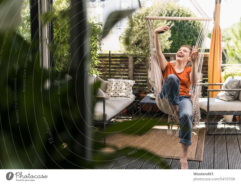Cheerful woman taking selfie with smart phone while sitting on swing in porch seen through doorway color image colour image Germany leisure activity