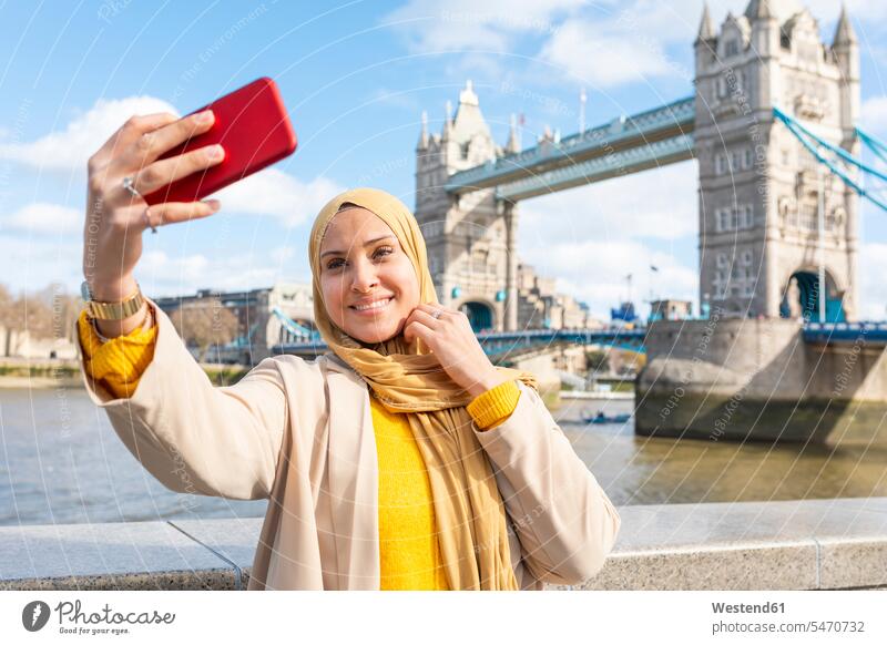 Portrait of smiling young woman taking selfie with smartphone in front of Tower Bridge, London, UK human human being human beings humans person persons