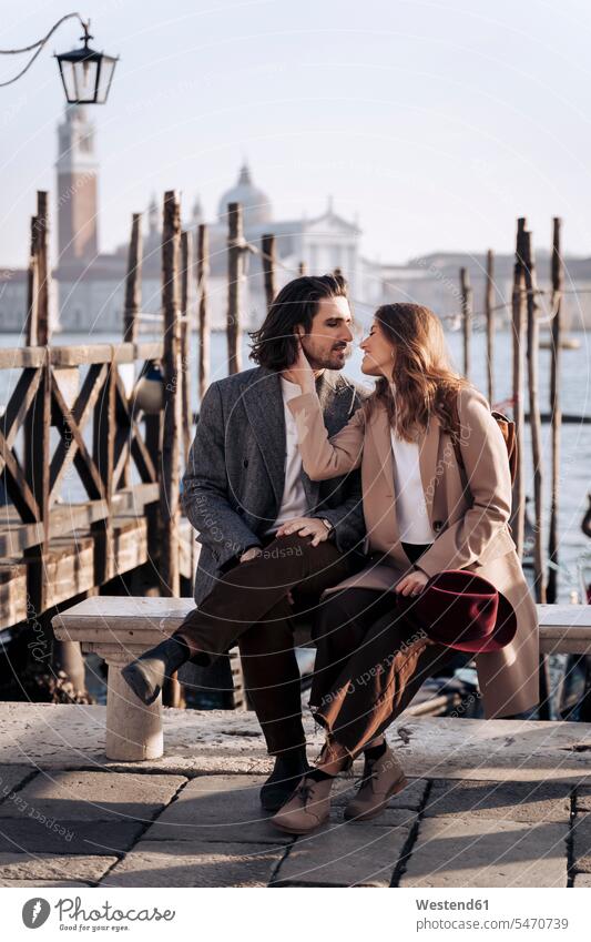 Young couple sitting at the waterfront in Venice, Italy hats coat coats jackets benches kiss kisses smile Seated relax relaxing relaxation delight enjoyment