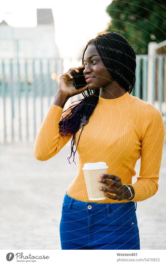 Smiling teenage girl with coffee cup looking away while talking on smart phone at street color image colour image outdoors location shots outdoor shot