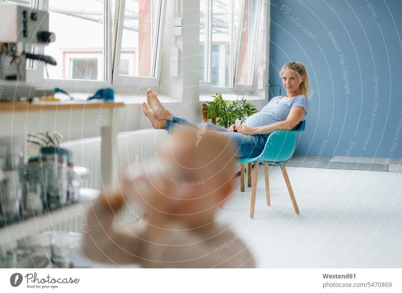 Pregnant woman sitting on a chair at the window with toddler in foreground Seated pregnant Pregnant Woman chairs females women windows Adults grown-ups grownups