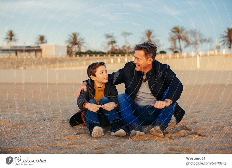 Smiling father embracing son on the beach beaches smiling smile sitting Seated sons manchild manchildren embrace Embracement hug hugging pa fathers daddy dads