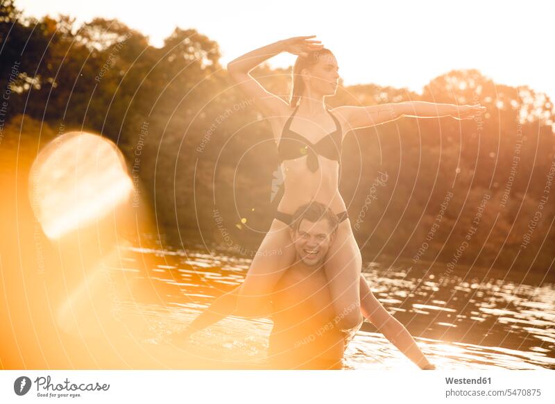 Young woman sitting on shoulders of young man standing in lake, enjoying the sun swim wear bikinis relax relaxing summer time summertime summery delight
