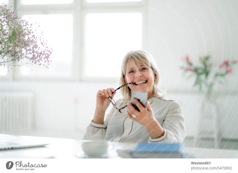 Portrait of happy mature businesswoman holding cell phone at desk happiness businesswomen business woman business women desks mobile phone mobiles mobile phones
