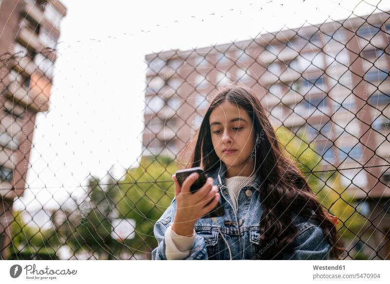Low angle view of teenage girl using smart phone while standing against chainlink fence color image colour image outdoors location shots outdoor shot