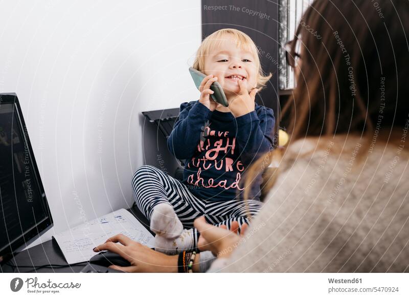 Mother working at home with little daughter sitting on desk holding cell phone mother mommy mothers ma mummy mama Seated daughters At Work mobile phone mobiles