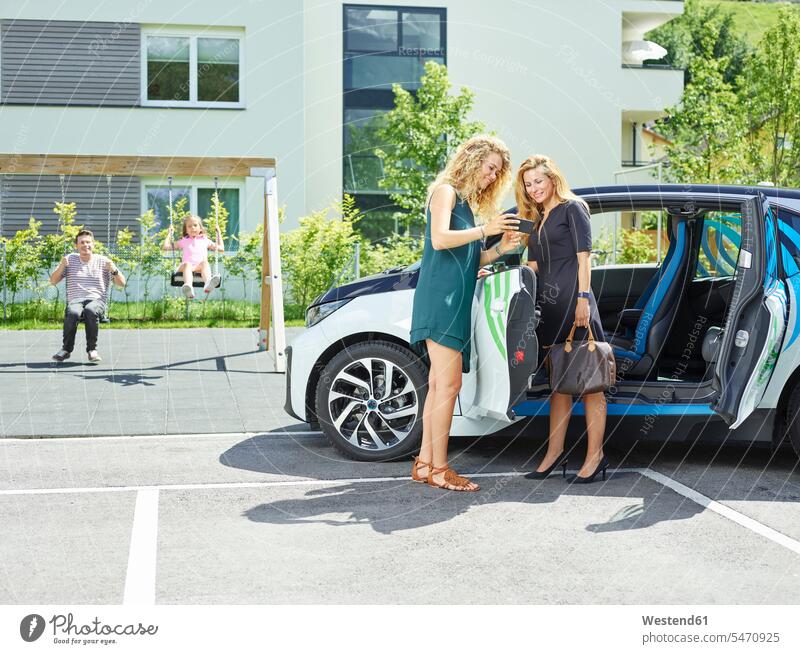 Two women with cell phone at electric car and man and girl in background automobile Auto cars motorcars Automobiles woman females mobile phone mobile phones