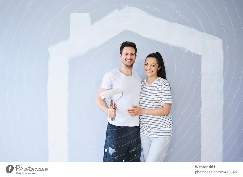 Portrait of happy couple painting in new apartment with house shape on wall shapes portrait portraits happiness twosomes partnership couples flat flats