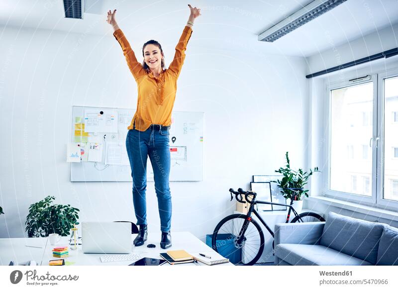 Happy young businesswoman cheering in office standing on desk happiness happy jubilate rejoicing rejoice exultation jubilating offices office room office rooms