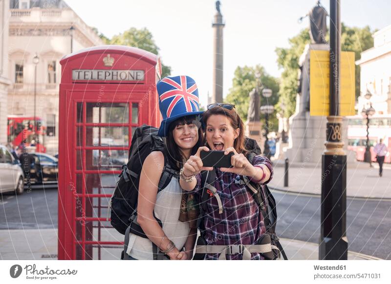 Cheerful woman taking selfie with friend wearing British flag hat against red telephone box in city color image colour image outdoors location shots