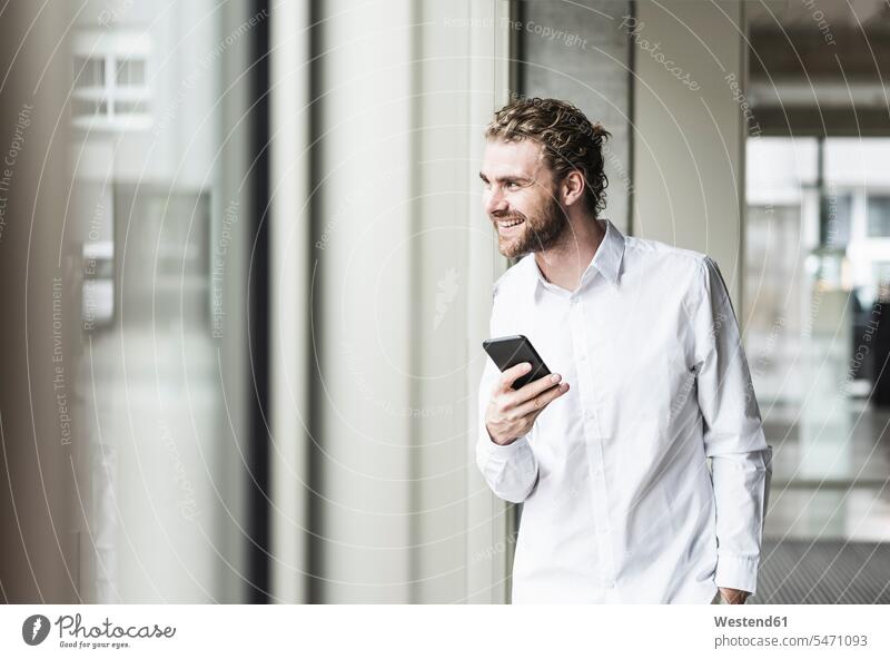Smiling young businessman holding cell phone looking out of window in office smiling smile men males offices office room office rooms windows mobile phone