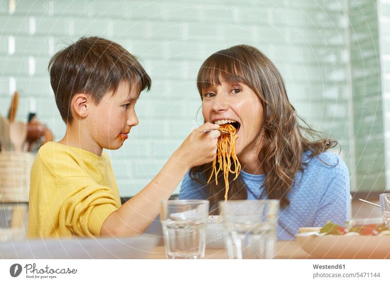 Son feeding mother with spaghetti in the kitchen dish dishes Plates smile Seated sit delight enjoyment Pleasant pleasure Cheerfulness exhilaration gaiety gay