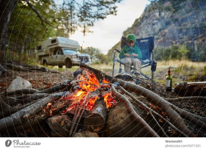 Chile, Santa Magda, Rio Maniguales, boy sitting at campfire in forest with camper van in background boys males Seated woods forests Camp Fire Campfire Bonfire