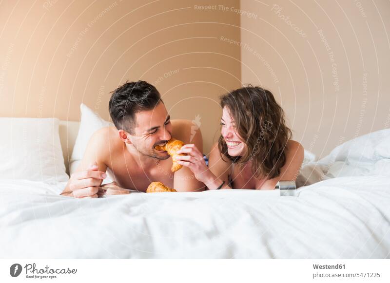 Happy young woman feeding croissant to man while lying in bed at home color image colour image indoors indoor shot indoor shots interior interior view Interiors