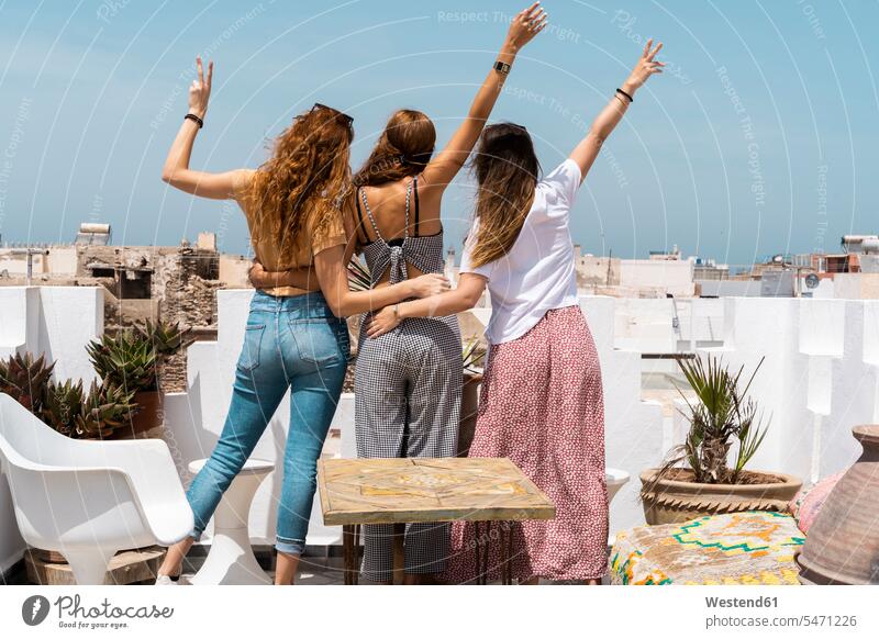 Back view of three young women standing on roof terrace showing victory signs, Essaouira, Morocco human human being human beings humans person persons