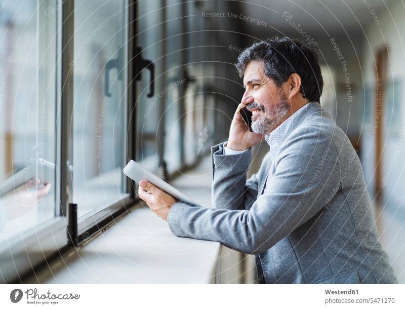Smiling male professor talking on phone while looking through window in corridor at university color image colour image day daylight shot daylight shots