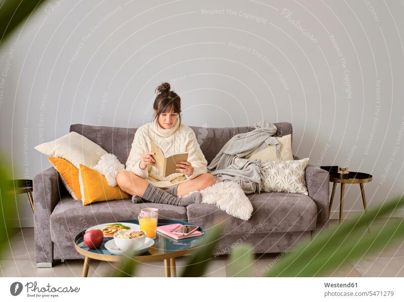 Woman reading book while sitting on sofa against wall at home during winter color image colour image Spain indoors indoor shot indoor shots interior