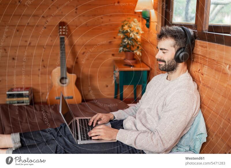 Smiling young man using laptop while listening music on bed in log cabin color image colour image outdoors location shots outdoor shot outdoor shots front view