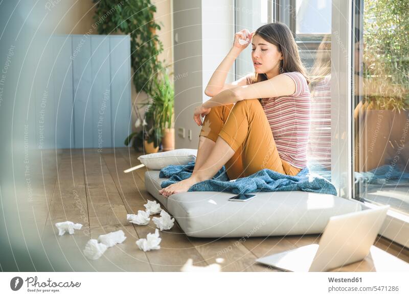 Sad young woman sitting at the window at home with laptop and tissues tissue handkerchief tissue handkerchiefs sad females women Seated Laptop Computers laptops