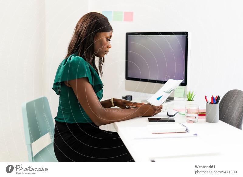 Businesswoman analyzing graph paper while sitting by desk at office color image colour image indoors indoor shot indoor shots interior interior view Interiors