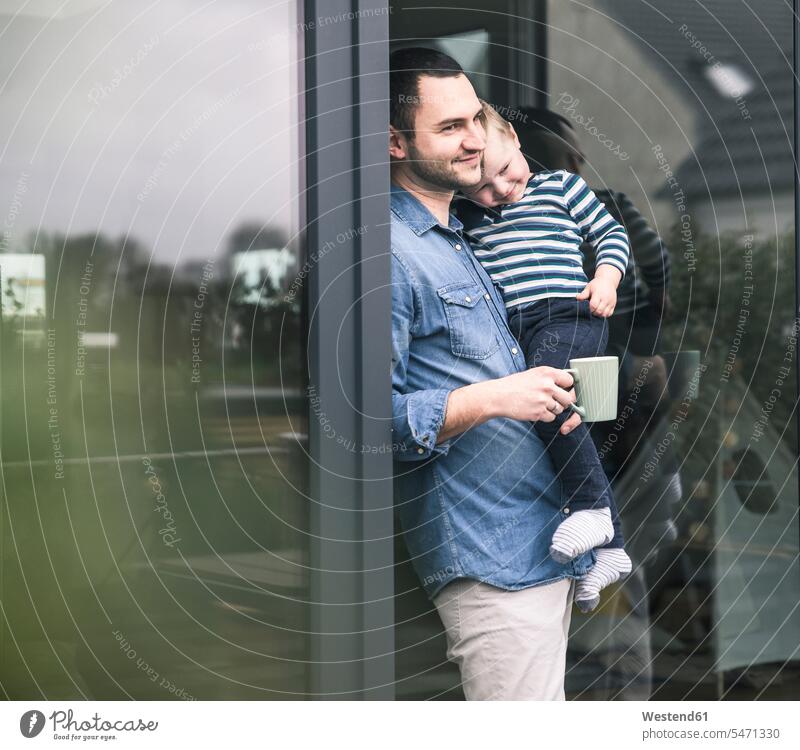 Father with cup of coffee carrying son at terrace door at home patio door French window father fathers daddy dads papa sons manchild manchildren Coffee Cup
