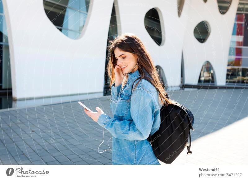 Spain, Barcelona, smiling young woman with backpack listening music with cell phone and earphones females women ear phone ear phones smile Smartphone iPhone