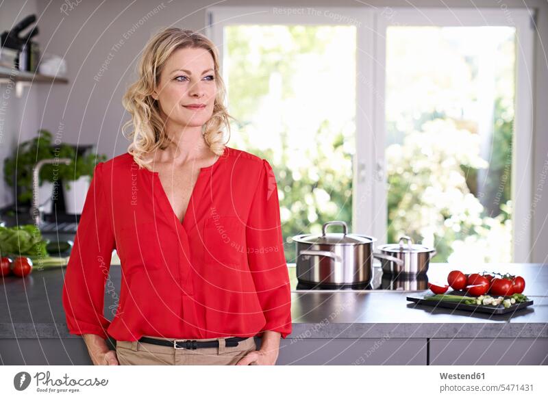Confident woman wearing red blouse standing in kitchen blouses confidence confident domestic kitchen kitchens females women colour colours Adults grown-ups