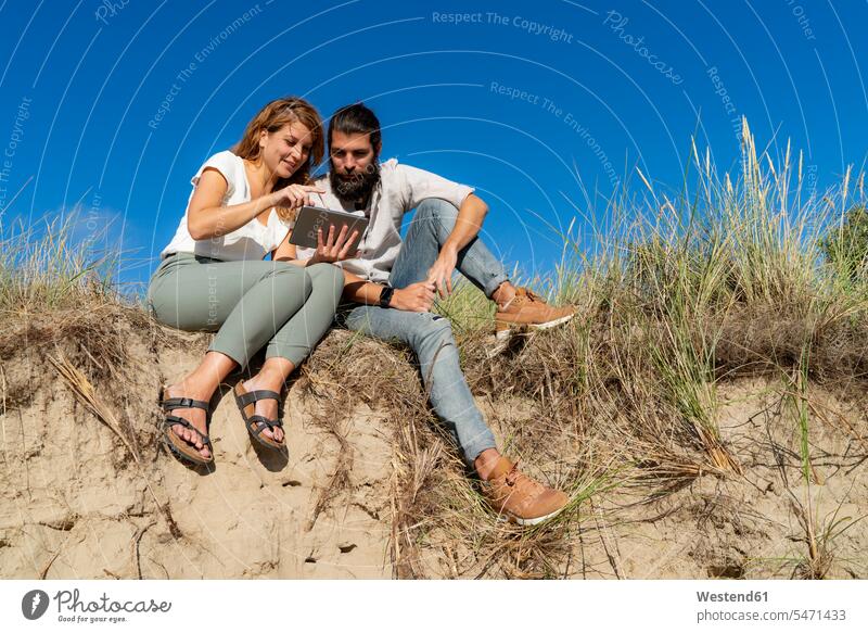 Young couple sitting on a dune in summer, using digital tablet recovering young couple young couples young twosome young twosomes summer time summery summertime