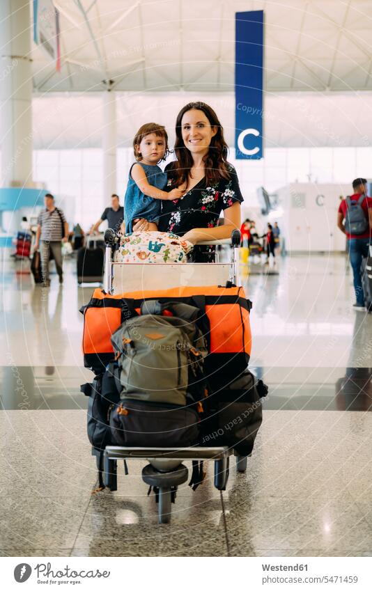 Mother holding a baby girl at the airport and pushing a trolley with the luggage standing smiling smile daughter daughters baggage cart luggage cart