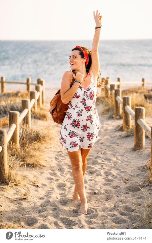 Young woman with raised arm walking at the beach in the evening fences back-pack back-packs backpacks rucksack rucksacks go going smile Late Evening summer time