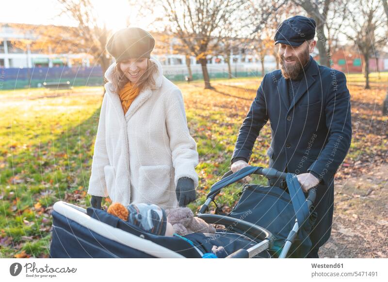 Happy couple with little son in a pushchair at park coat coats jackets caps hat hats baby carriage baby carriages prams stroller strollers smile delight