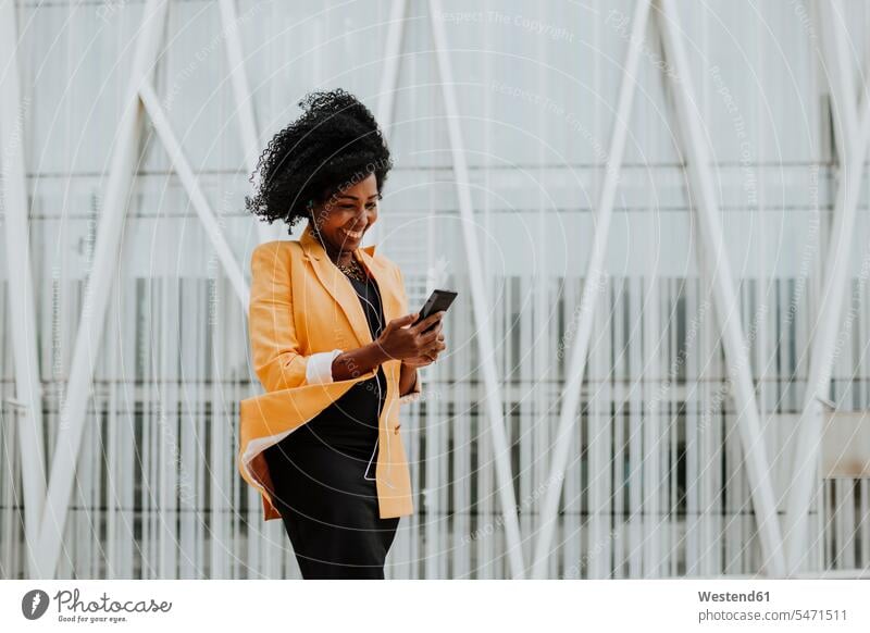 Smiling female entrepreneur using smart phone listening to music through earphones standing against building color image colour image outdoors location shots