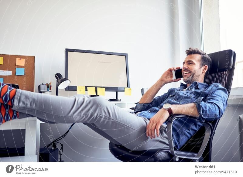 Relaxed man sitting at desk in office talking on cell phone on the phone call telephoning On The Telephone calling offices office room office rooms mobile phone