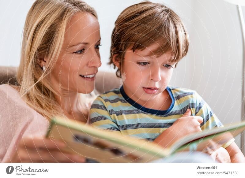 Mother and son reading a book together on couch at home books couches settee settees sofa sofas smile Seated sit relax relaxing relaxation delight enjoyment