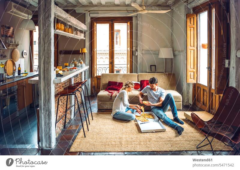 Young couple sitting on floor of living room and eating pizza from box boxes couches settee settees sofa sofas carpets rug rugs Seated at home free time
