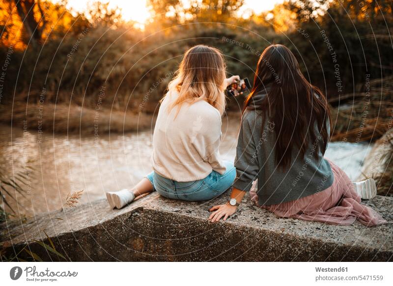 Sisters taking selfie while sitting on retaining wall in forest during sunset color image colour image outdoors location shots outdoor shot outdoor shots
