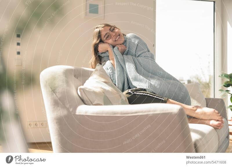 Happy young woman with woolen blanket on couch Germany flat flats apartment apartments Wellbeing Well-Being Well Being lifestyle life styles comfortable