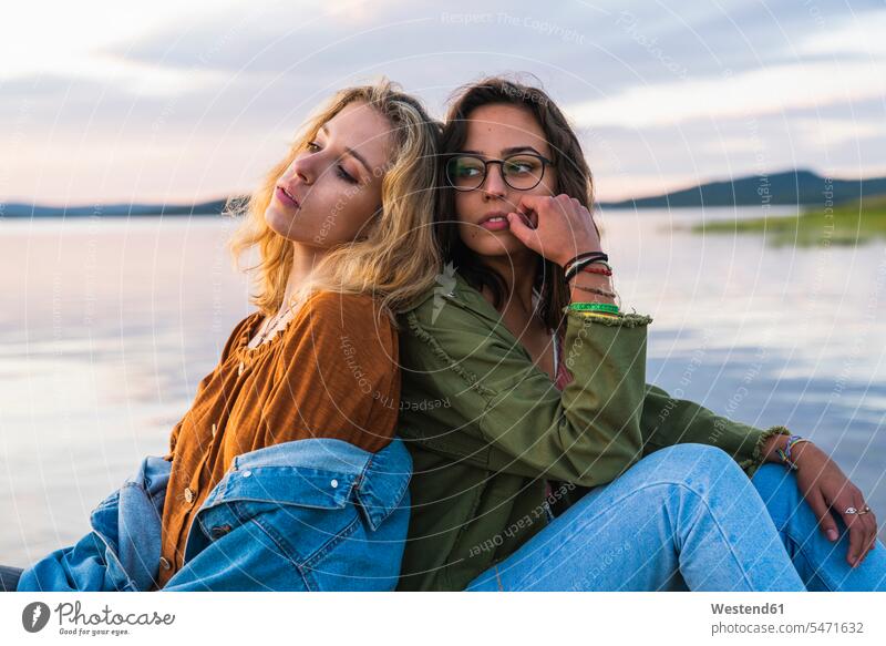 Finland, Lapland, two young women sitting back to back at the lakeside at twilight woman females girlfriend Girlfriends girl friend girl friends Seated water