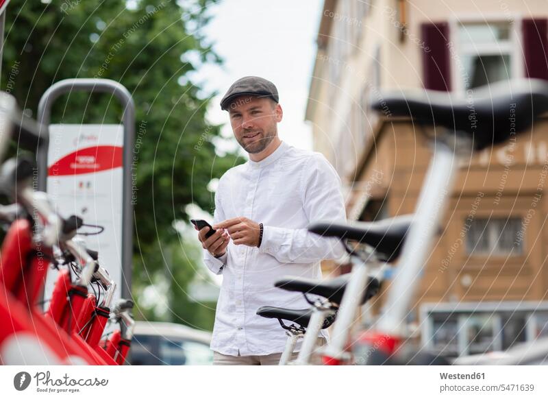 Smiling man renting bicycle through smart phone at parking station in city color image colour image Germany outdoors location shots outdoor shot outdoor shots