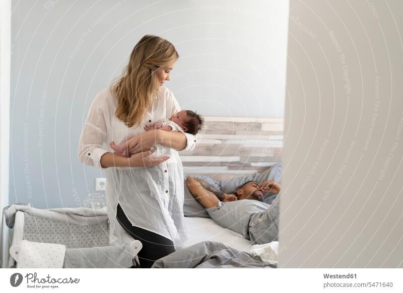 Mother holding her newborn baby in bedroom with father sleeping in background family with one child focus on foreground Focus In The Foreground