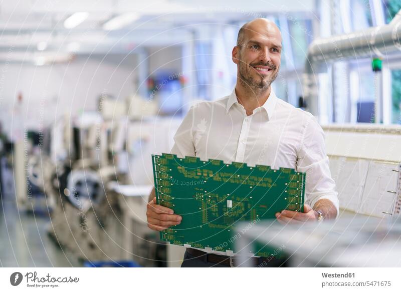 Smiling male technician holding large circuit board while looking away at factory color image colour image indoors indoor shot indoor shots interior