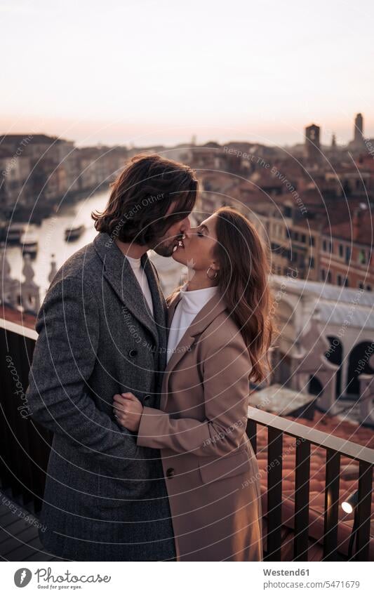 Affectionate young couple kissing on a balcony above the city of Venice, Italy coat coats jackets kisses in the evening Late Evening enjoy enjoyment indulgence