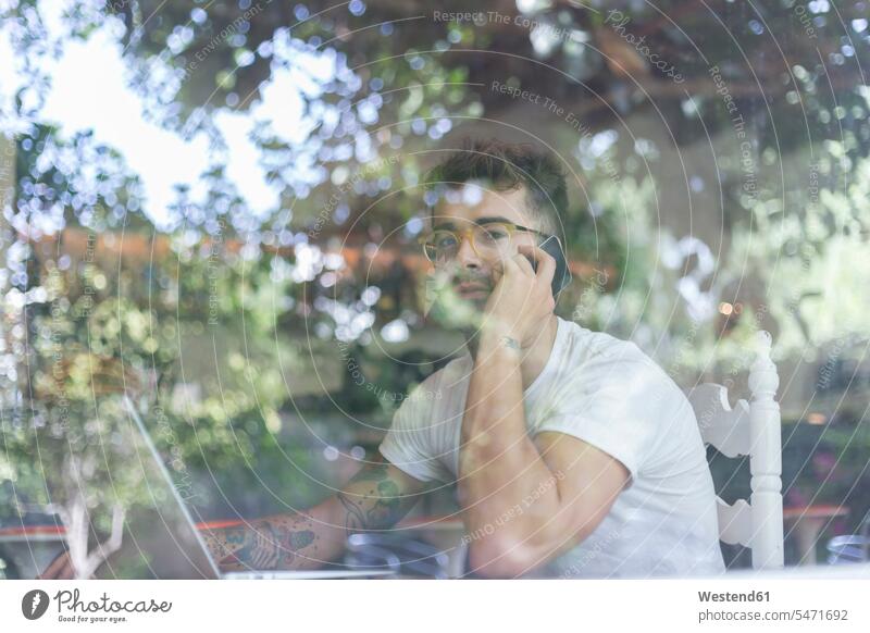 Young man with laptop on cell phone behind windowpane of a cafe windows panes window glass window glasses Window Pane windowpanes T- Shirt t-shirts tee-shirt