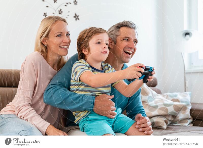 Happy parents with son playing video game on couch at home couches settee settees sofa sofas hold smile Seated sit relax relaxing relaxation delight enjoyment